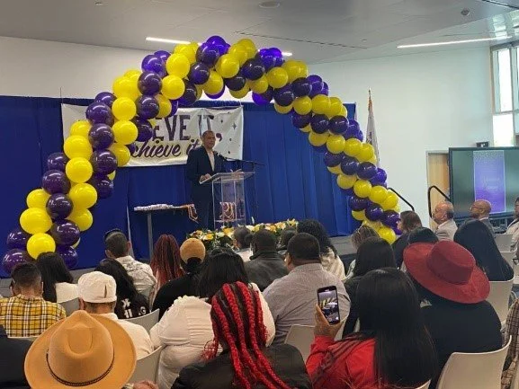 A person speaking at a podium on a stage decorated with purple and yellow balloon arch, with an audience seated and some taking photos, in a conference or event setting.