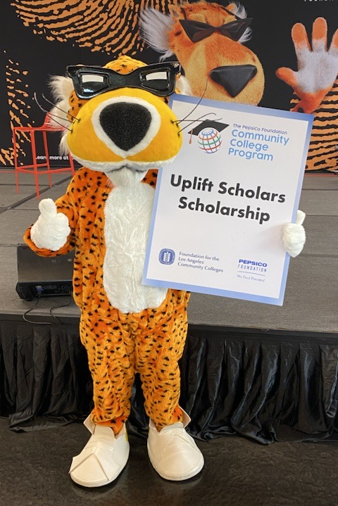 Costumed cheetah mascot standing on stage, wearing glasses, holding a sign that says "Uplift Scholars Scholarship," with a black table and colorful large animal face backdrop in the background.