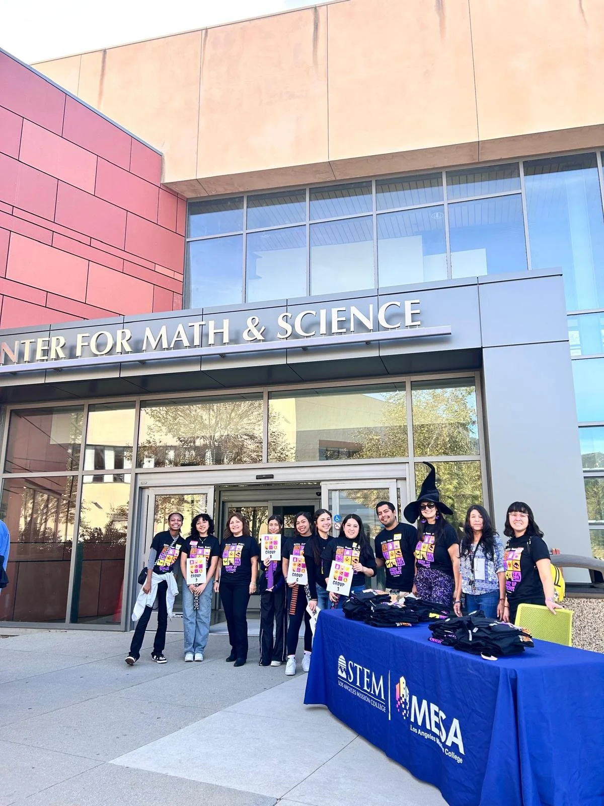 Group of people standing outside the Center for Math & Science building with a table displaying STEM and MESA banners.