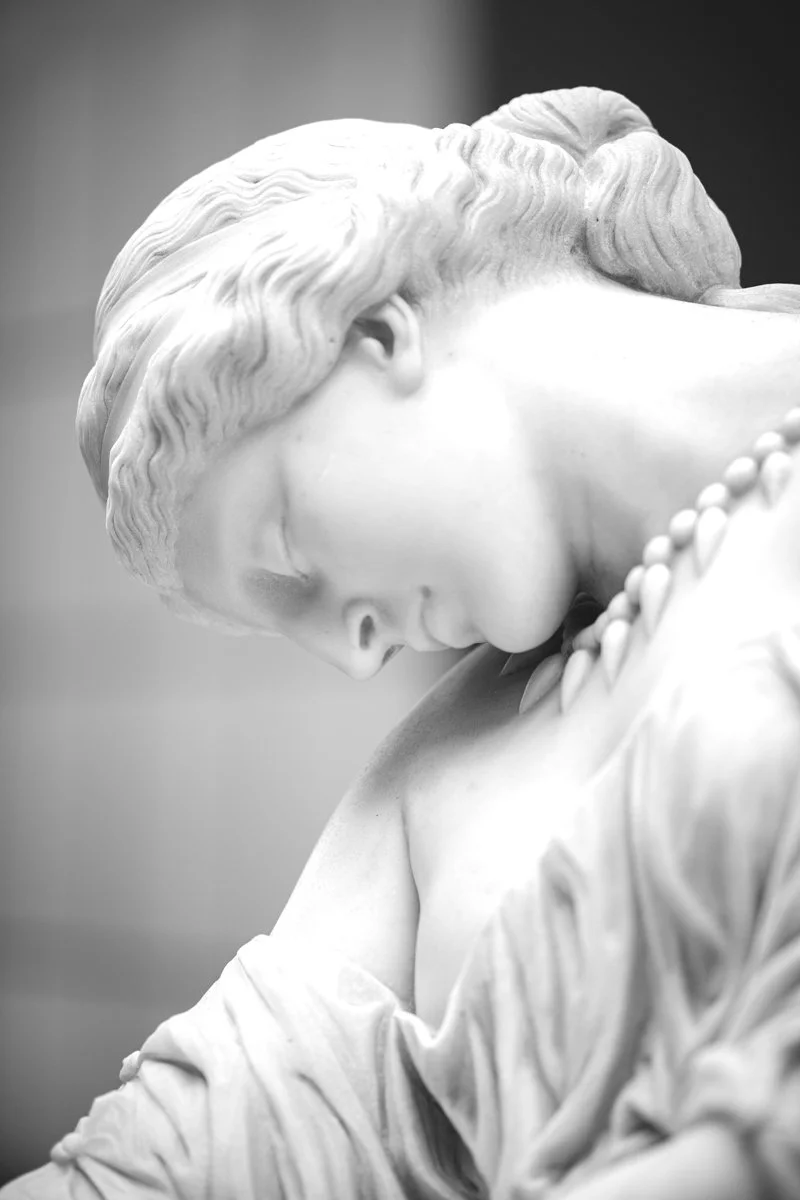 A close-up black and white photo of a classical marble sculpture of a woman with her head bowed and eyes closed, with detailed hair and draped clothing.