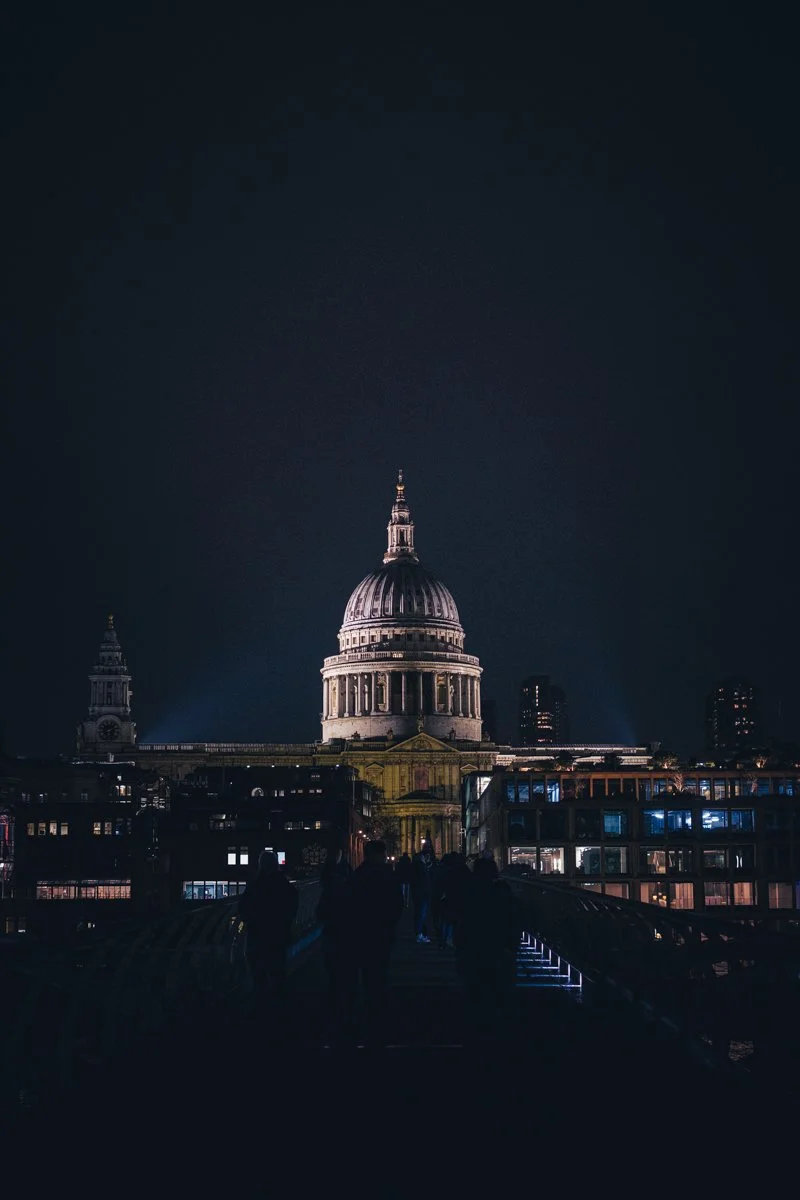 Night view of St. Paul's Cathedral in London illuminated against the dark sky with silhouettes of people in the foreground.