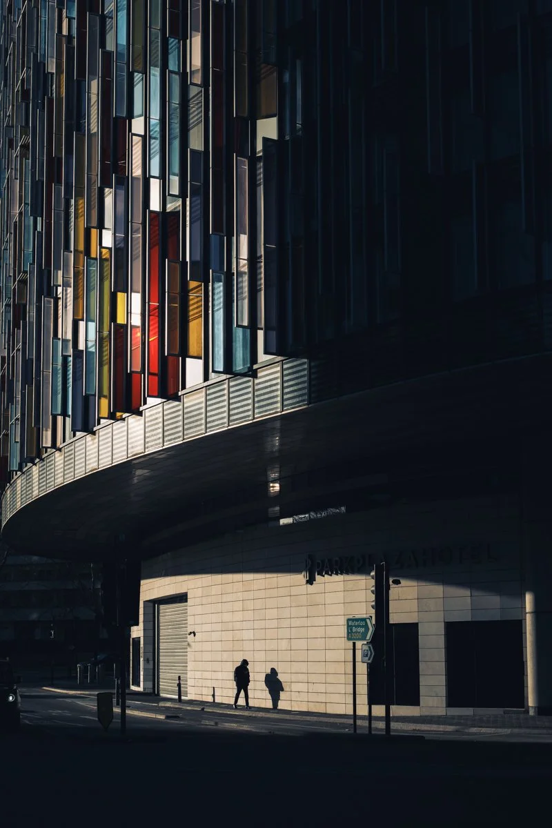A modern building with colorful glass windows and a curved facade, featuring two people walking on the sidewalk beneath it.
