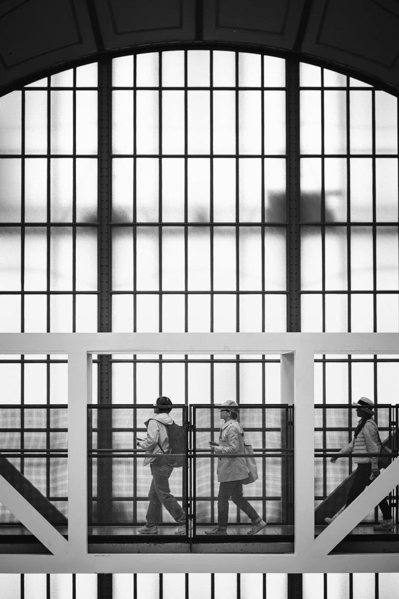 Three people walking on an elevated enclosed walkway inside a building with large arched window wall.
