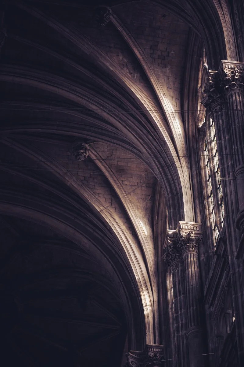 Interior view of a Gothic cathedral showing pointed arches, tall columns, and large stained glass windows with sunlight streaming through.