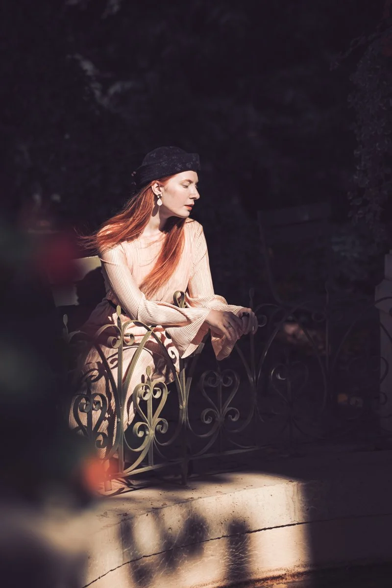 A woman with long red hair, wearing a black hat and earrings, sitting on a decorative wrought iron bench in a shadowy outdoor setting.