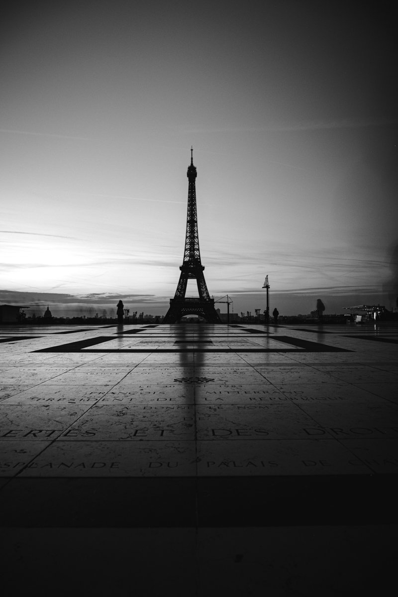Black and white photo of the Eiffel Tower in Paris, France, taken at sunset with a few people walking nearby.