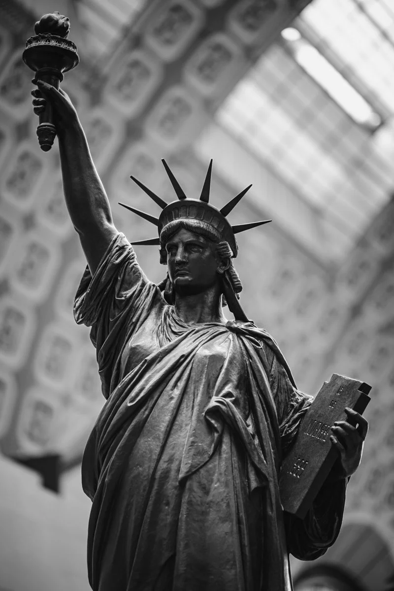 Black and white photograph of the Statue of Liberty holding a torch in her right hand and a tablet in her left hand. The background shows the interior of a building with decorative arches and a glass ceiling.