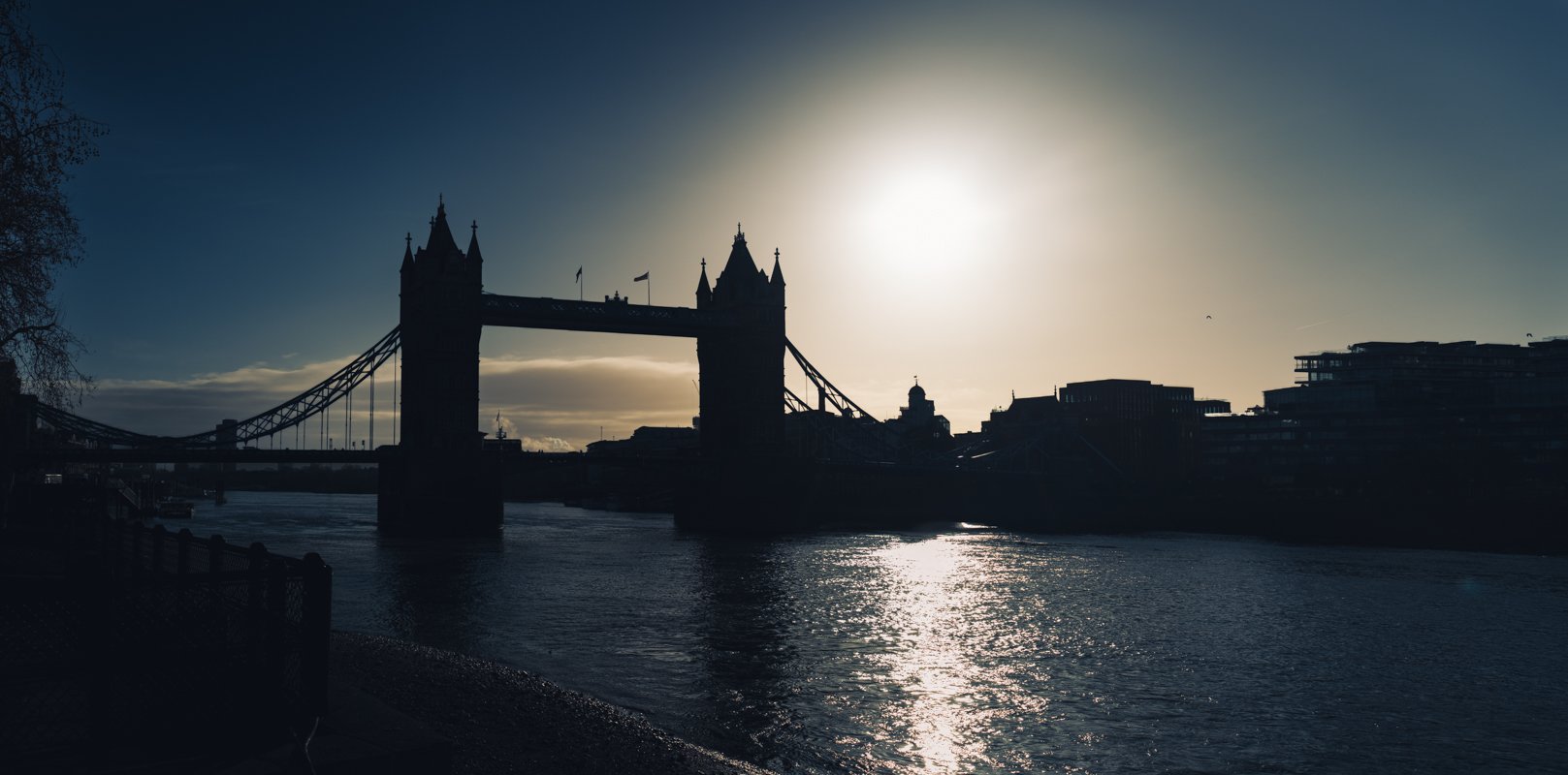Silhouette of Tower Bridge over the River Thames with the sun setting behind, creating a reflective glow on the water.