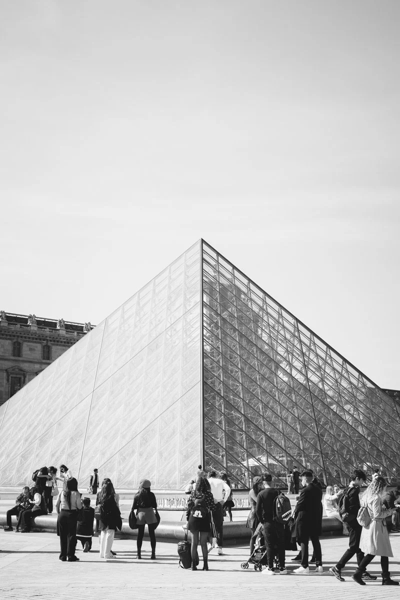 The Louvre Pyramid in Paris, made of glass, surrounded by people walking and sitting.