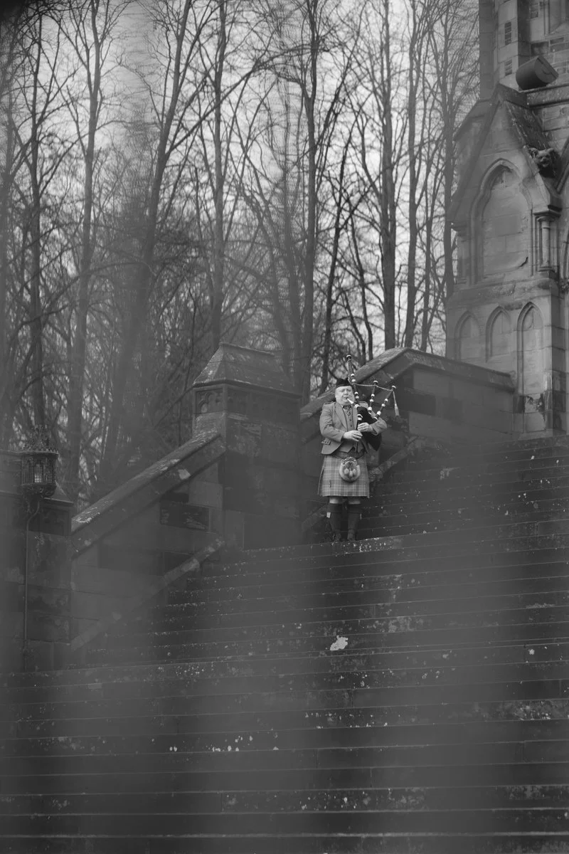 A man playing bagpipes on outdoor stone steps near a historic building and leafless trees in winter.