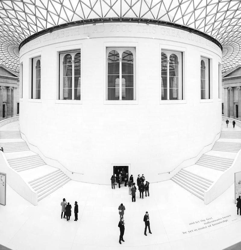 Interior view of the Large Round Reading Room at the British Museum, with white walls, large arched windows, and people walking around.
