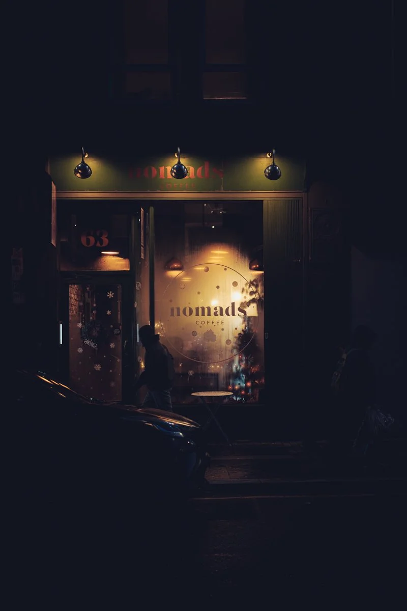Night time view of a coffee shop called 'Nomads Coffee' with a large illuminated window and door, and a person walking by in front of the shop.