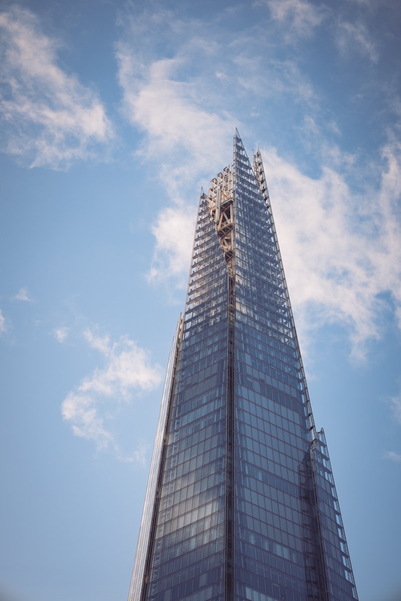 A tall modern glass skyscraper with a pointed top against a blue sky with scattered clouds.