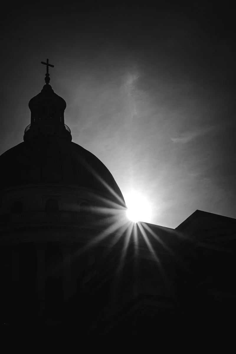 A church with a dome and cross, silhouetted against the sun in a black and white photo.