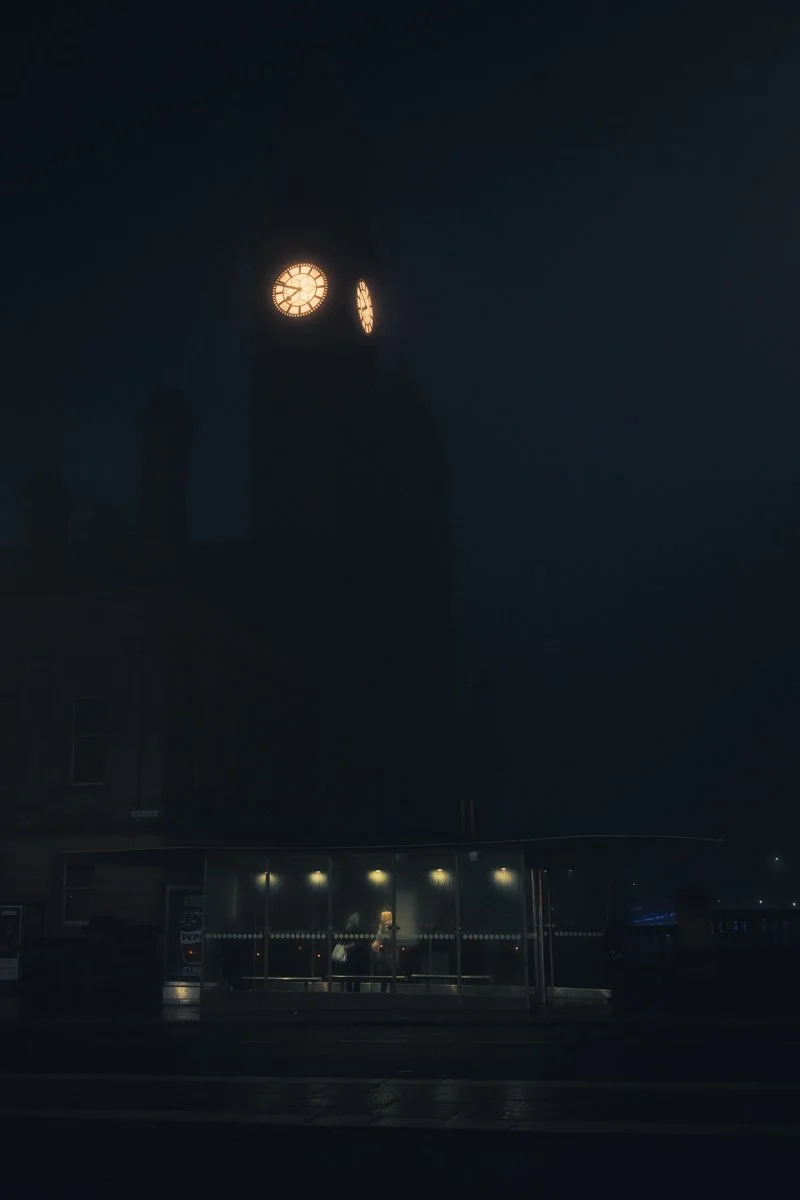 A clock tower lit up at night with visible clock faces, and a bus stop shelter with people waiting inside, illuminated against the dark sky.