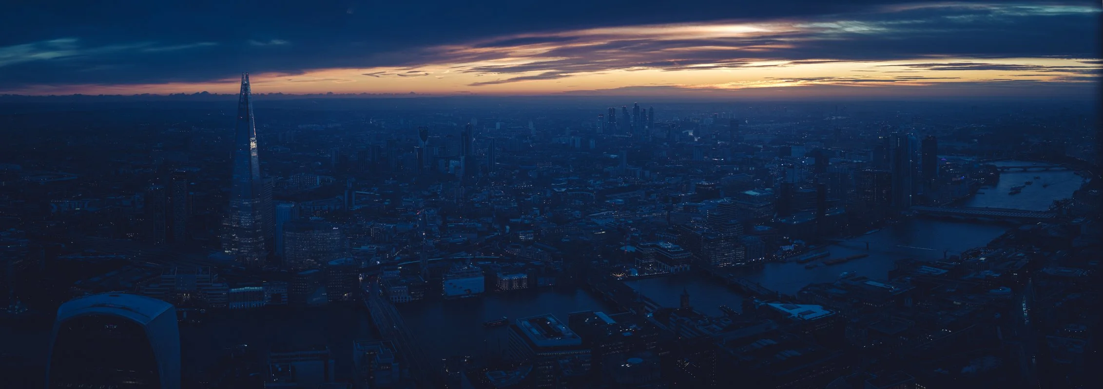 Nighttime aerial view of a city skyline, with tall skyscrapers, a river, and bridges, under a dark sky with some light on the horizon.