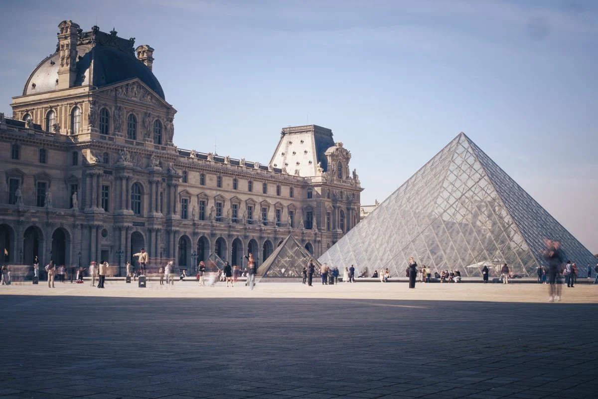 The Louvre Museum with its historic building and glass pyramid entrance in Paris, France, on a clear day with visitors walking around the courtyard.