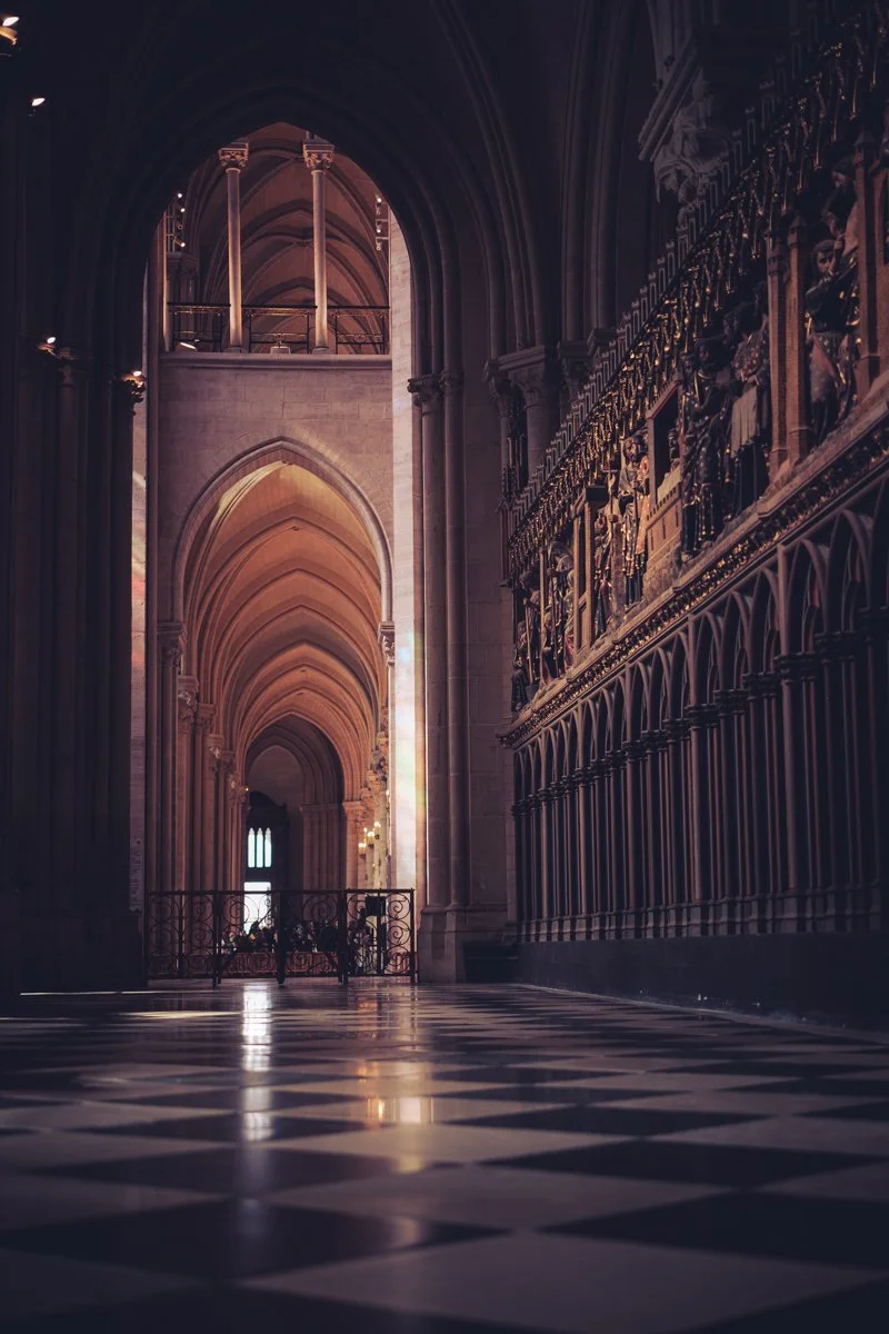 Interior of a Gothic-style cathedral with tall pointed arches, intricate decorative sculptures on the right wall, and a checkered black and white floor. Sunlight filters through a stained glass window at the end.