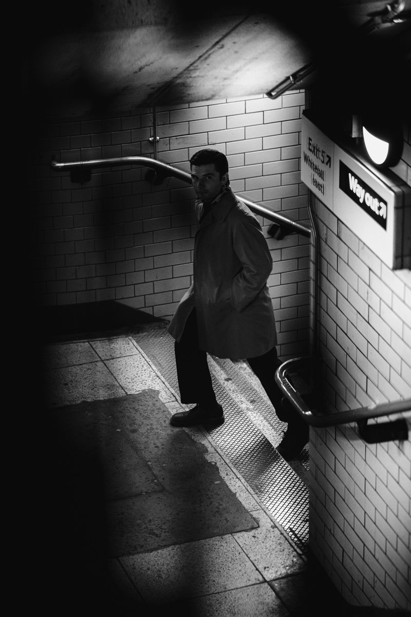 A man in a trench coat standing on a subway station platform, looking towards the camera, with tiled walls and illuminated exit signs.