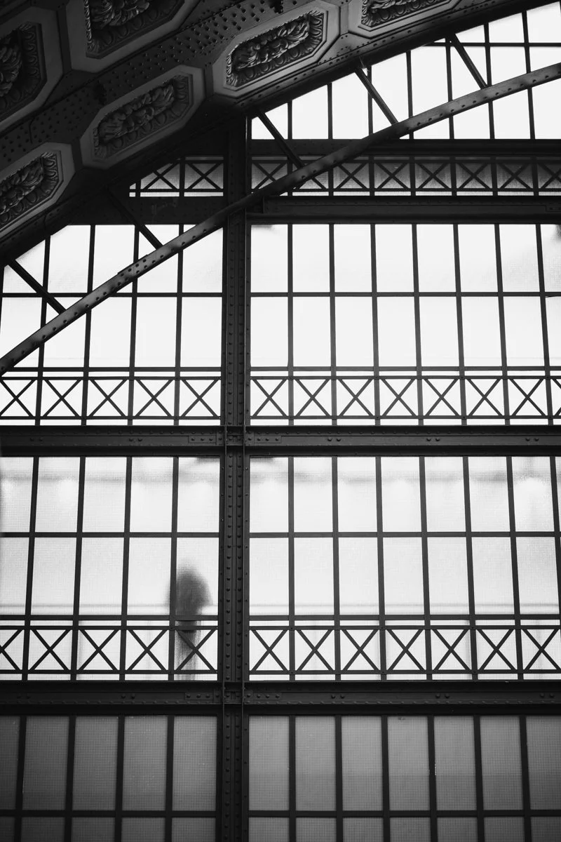 Black-and-white interior view of a large industrial-style building or train station with metal framework and glass windows. A blurred person is visible behind the windows.