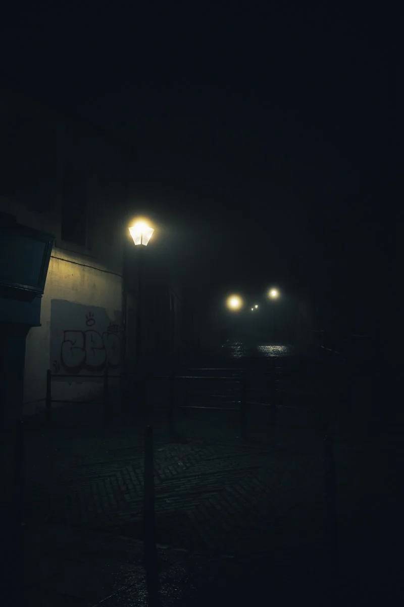 A dark, foggy street at night with streetlights illuminating parts of the street and a wall with graffiti.