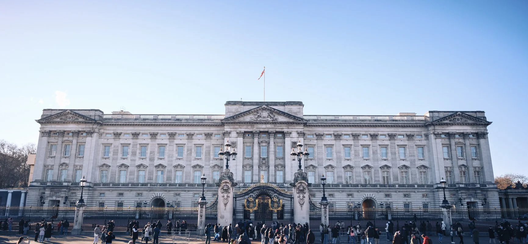Front view of a large historic white building with many windows, black wrought iron gate, and people walking in front, under a clear blue sky.