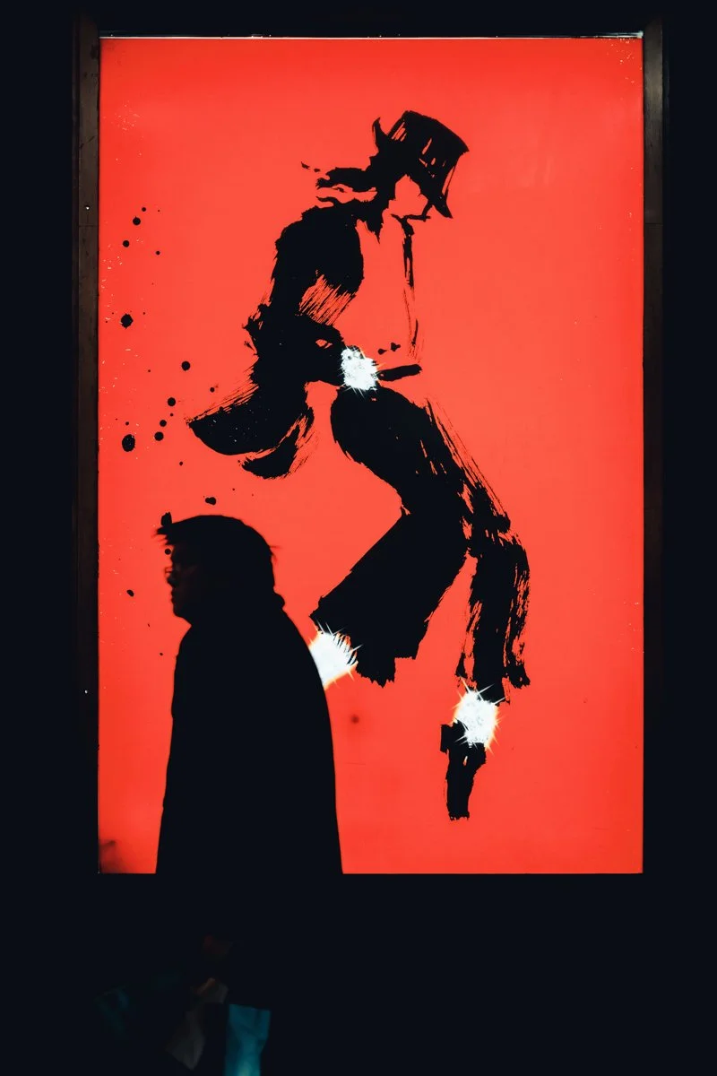 Silhouette of a man in front of a red background with black graffiti art of a figure with a hat, along with white paint splatters.