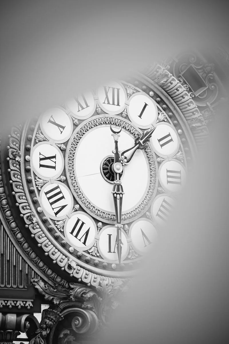 A black and white close-up of a vintage clock showing 12:07, with ornate decorative elements around the clock face.