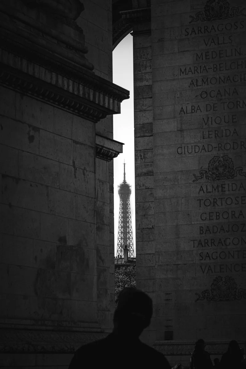Black and white photo of a person with glasses standing in front of a stone structure with inscriptions, framing a view of the Eiffel Tower in Paris.