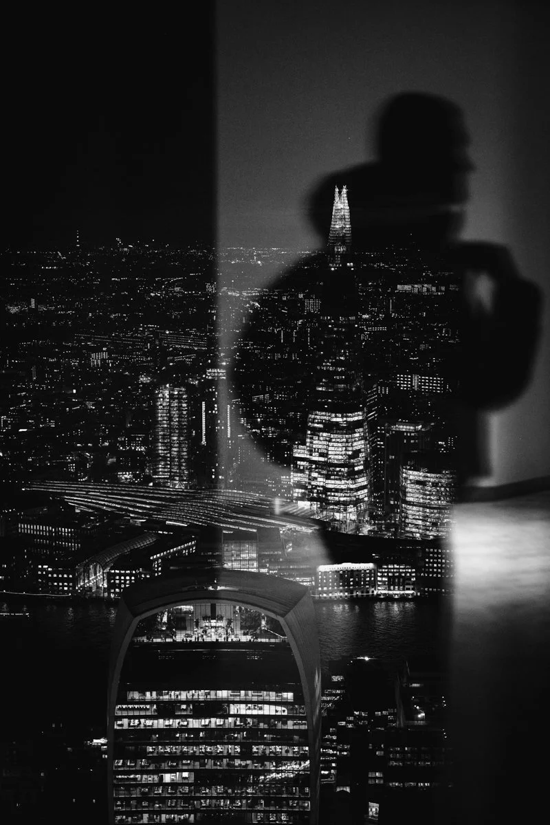 A black and white photo of a city skyline at night seen through a reflective glass, with a person's silhouette visible in the reflection.