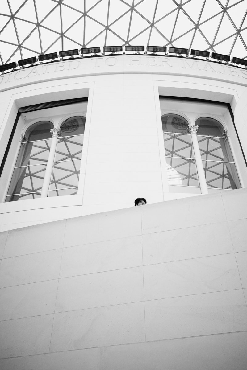 Black and white photo of a modern building interior, showing a person partially visible behind a wall with large arched windows, decorative columns, and a geometric glass ceiling.