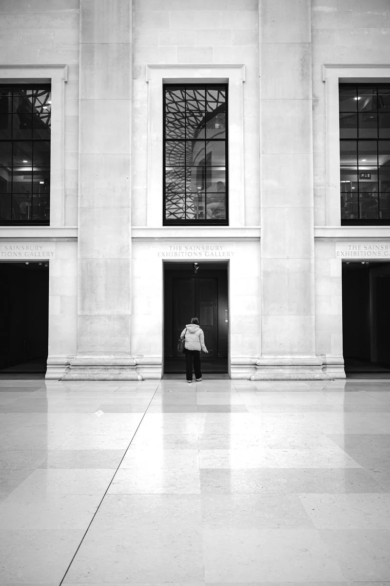 A person standing in front of a doorway inside an indoor gallery space with large windows and architectural details.
