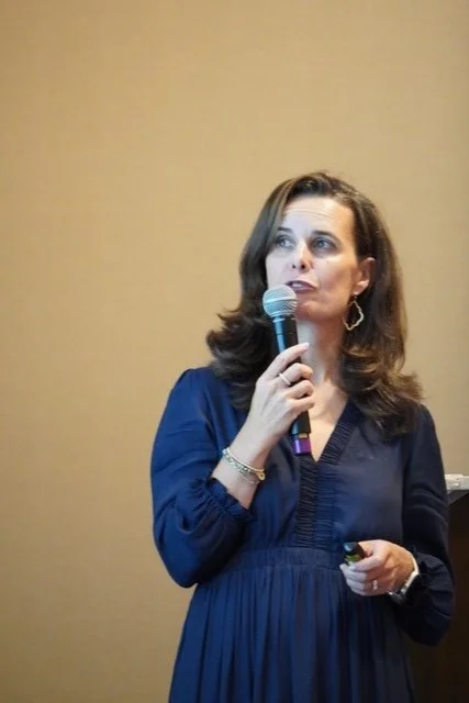 A woman with long dark hair wearing a blue dress holding a microphone and speaking.