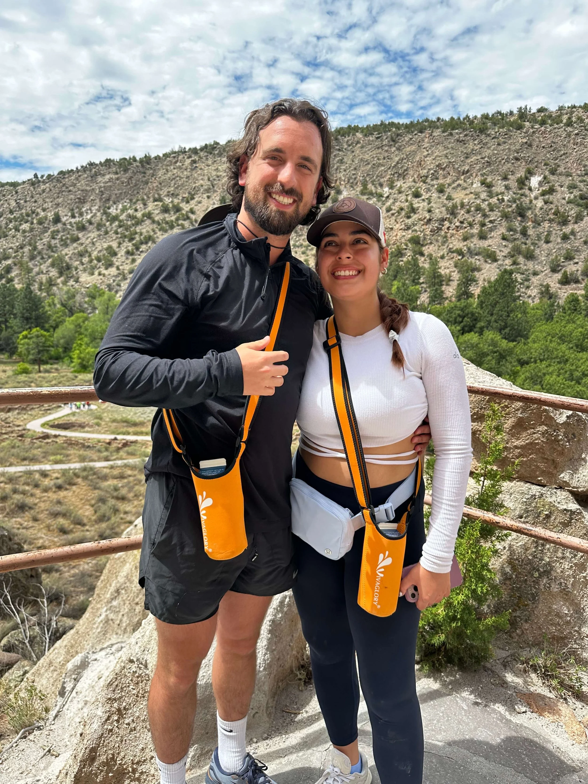 Two people posing outdoors, both wearing athletic clothing and carrying orange water bottle holders, with a rocky landscape in the background.