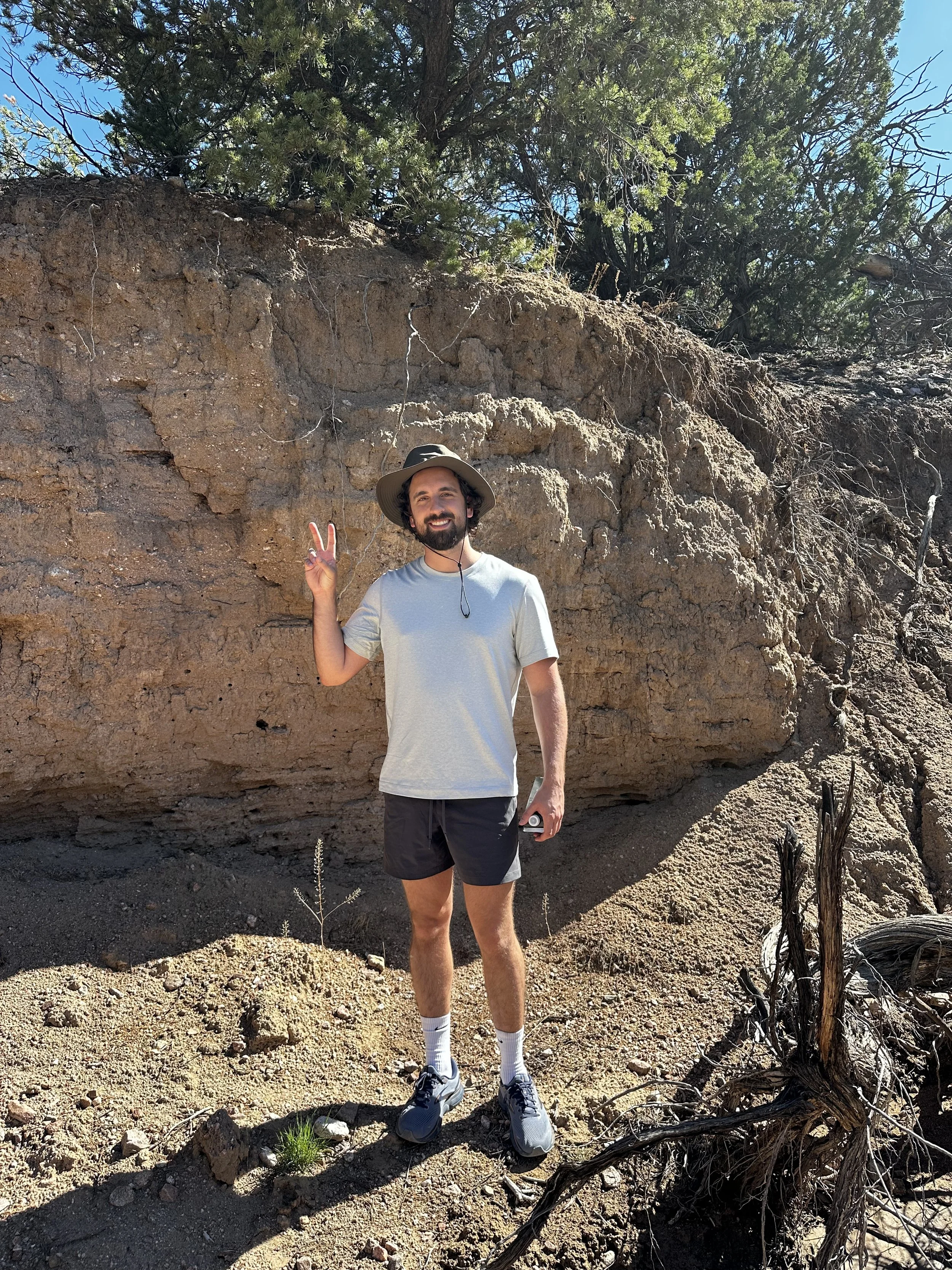 Man in gray shirt and shorts standing outdoors on rocky terrain with trees in the background, making a peace sign.