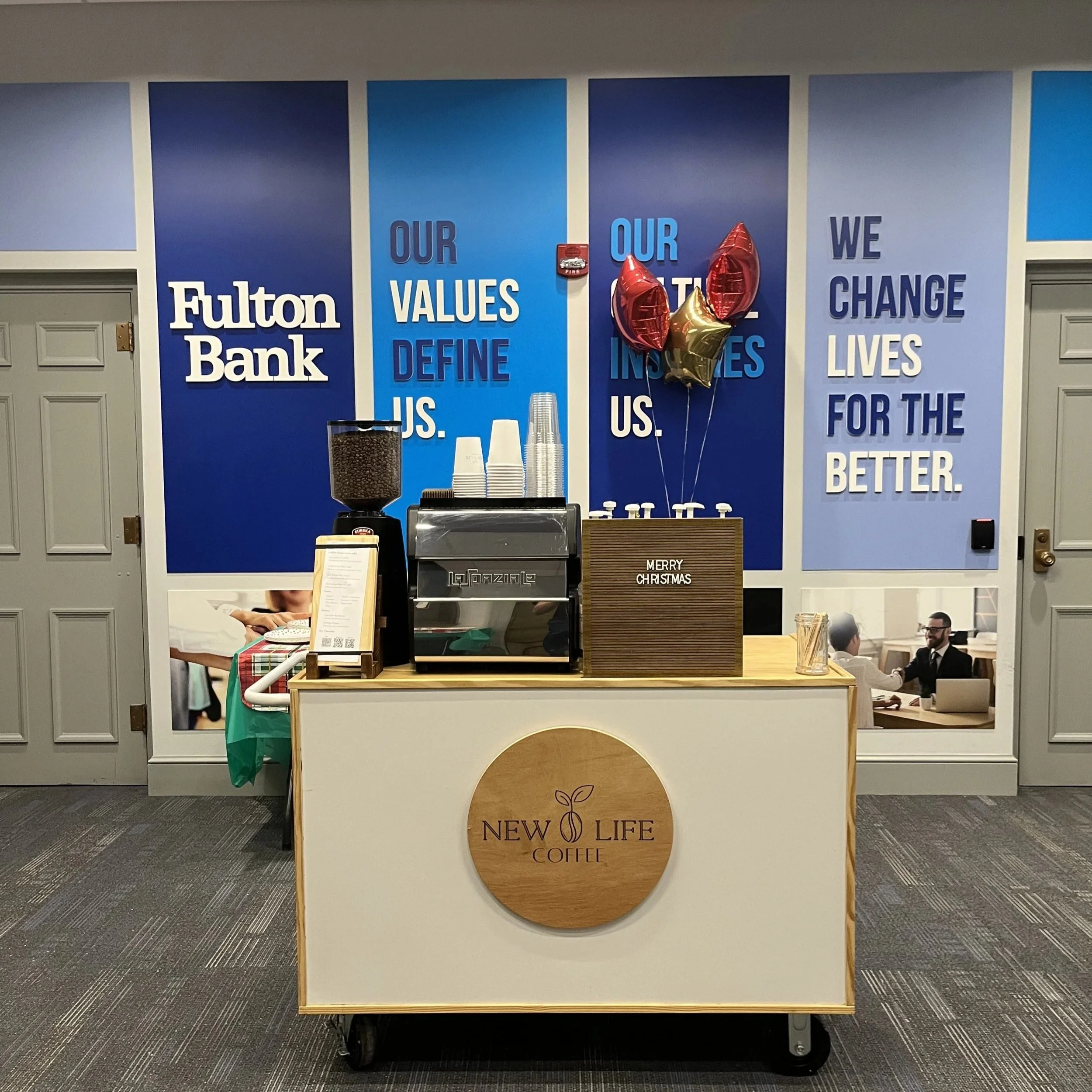 Coffee kiosk in an office with a Fulton Bank sign on the wall, decorated with balloons and holiday greeting sign, inside a corporate space.
