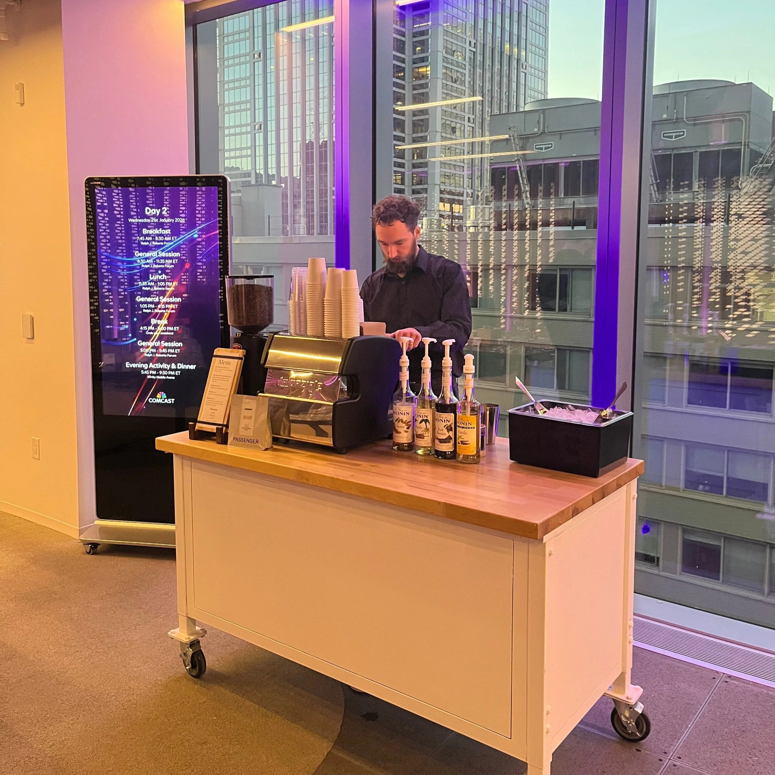 A barista standing behind a wooden cart with bottles of alcohol, an espresso machine, and cups, inside a high-rise building with large windows showing city buildings and purple lighting reflections.