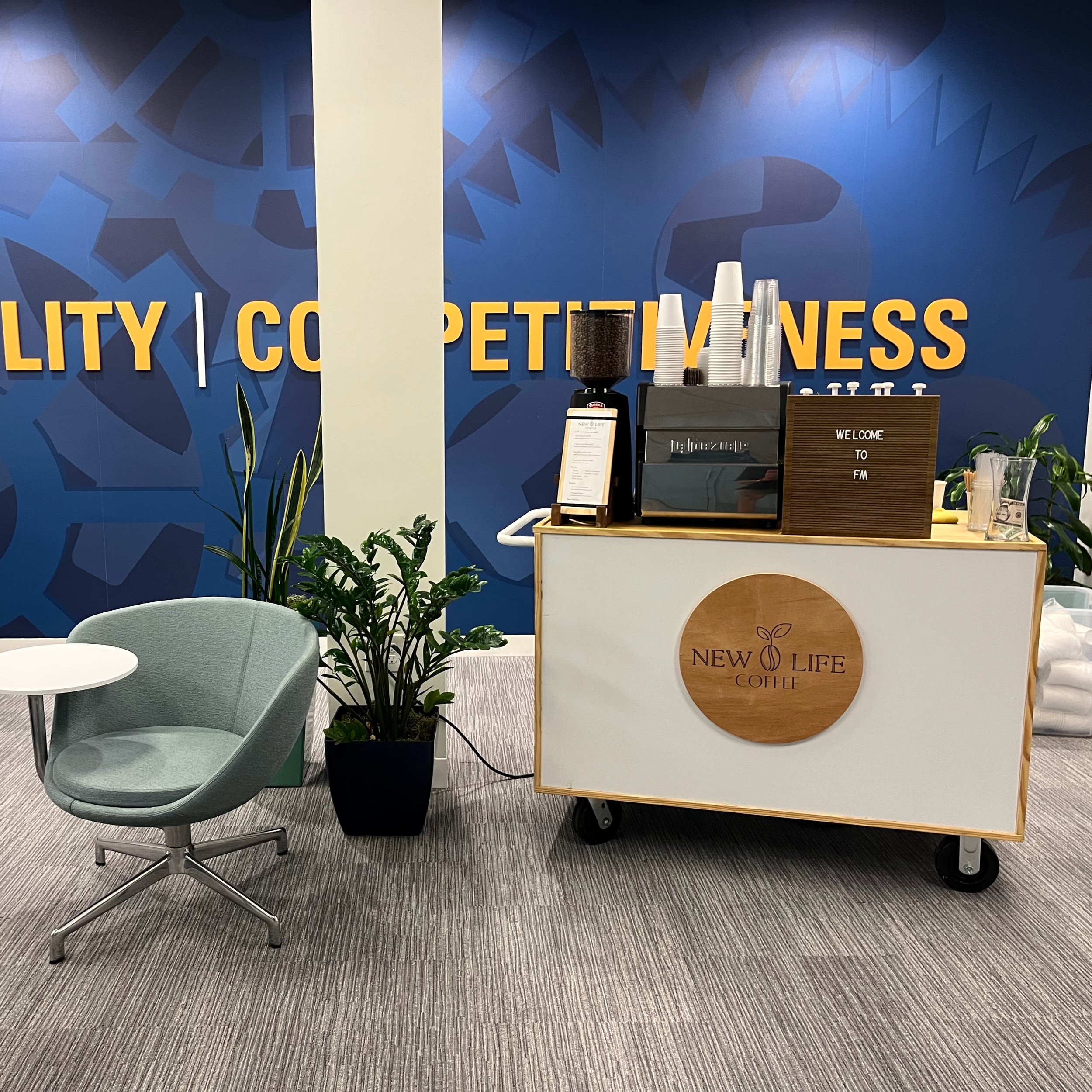 Coffee stand with a wooden sign reading 'New Life Coffee', coffee machine, cups, and plants in an office space with a blue wall that has the words 'Quality | Competitiveness'.