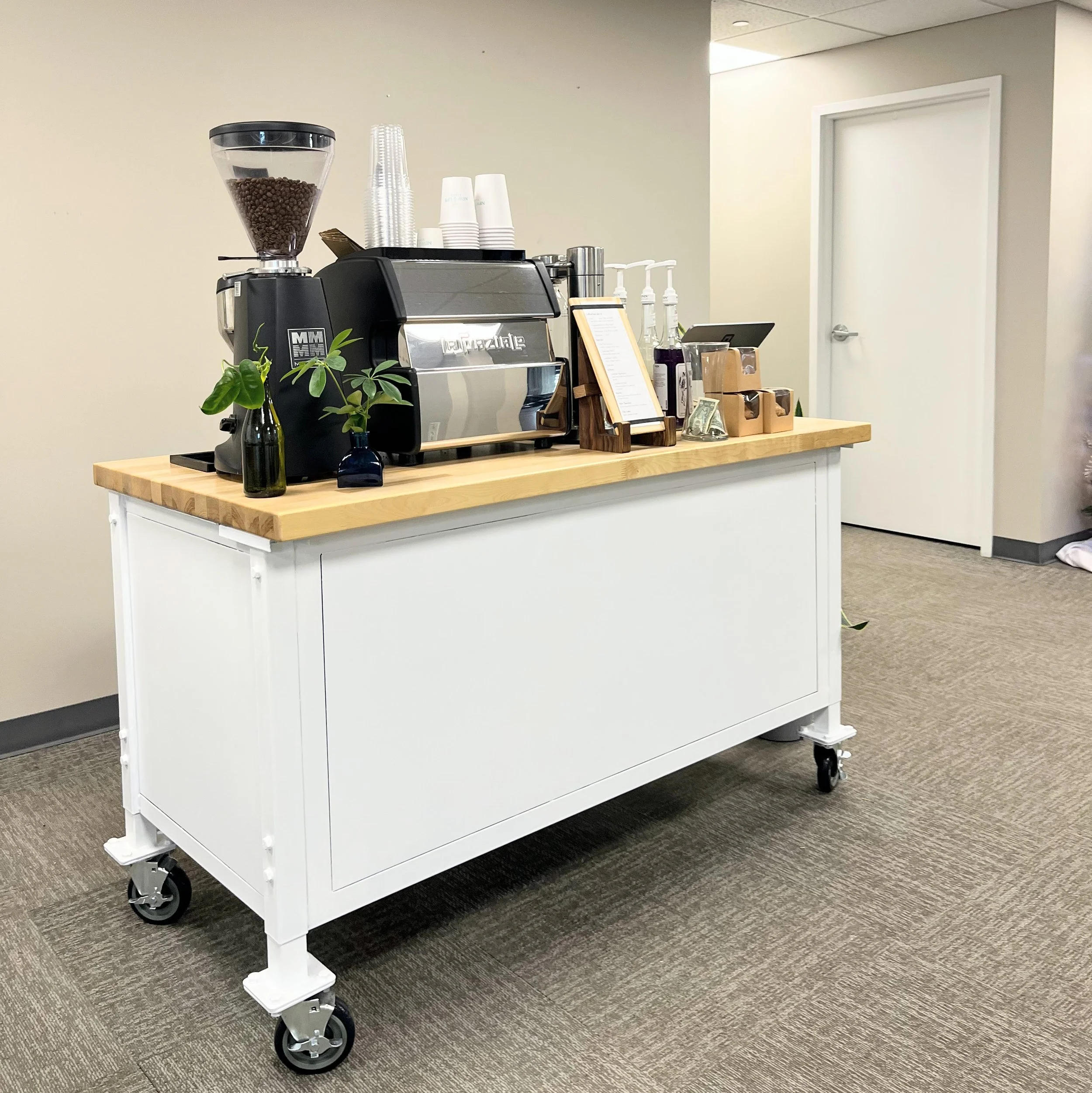 A mobile coffee station with a commercial espresso machine, a coffee grinder, cups, and small vases with green plants on a wooden table in a room with beige walls and gray carpeting.