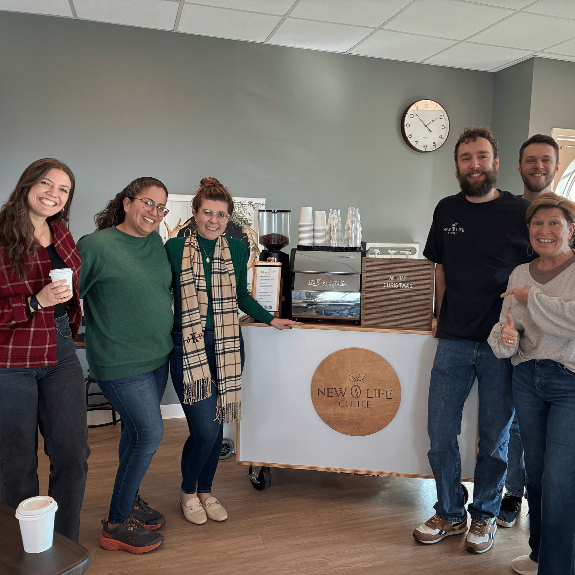 Group of six people standing behind a coffee counter with a coffee machine, cups, and a sign that says "Merry Christmas", smiling at the camera.