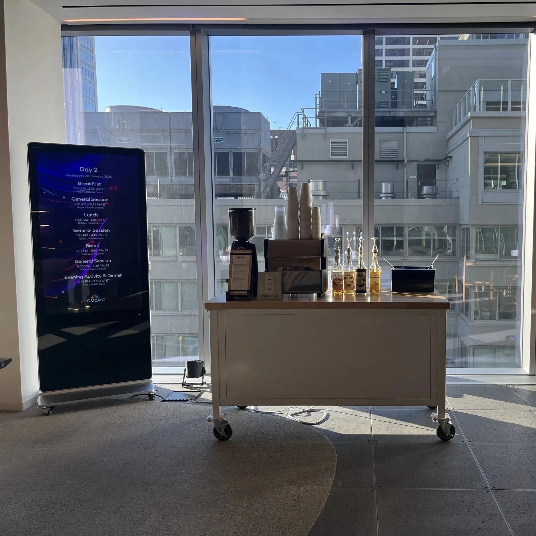 Coffee station with cups, bottles of syrup, and a menu sign in front of a large window showing city buildings outside.