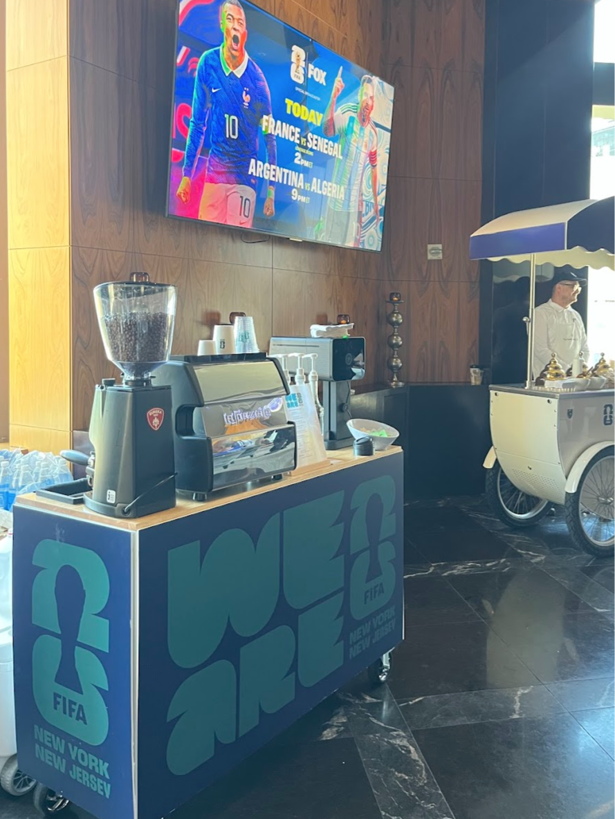A FIFA-branded beverage cart and coffee station in a modern interior, with a large TV screen showing a soccer match broadcast, featuring a soccer player and upcoming game details.