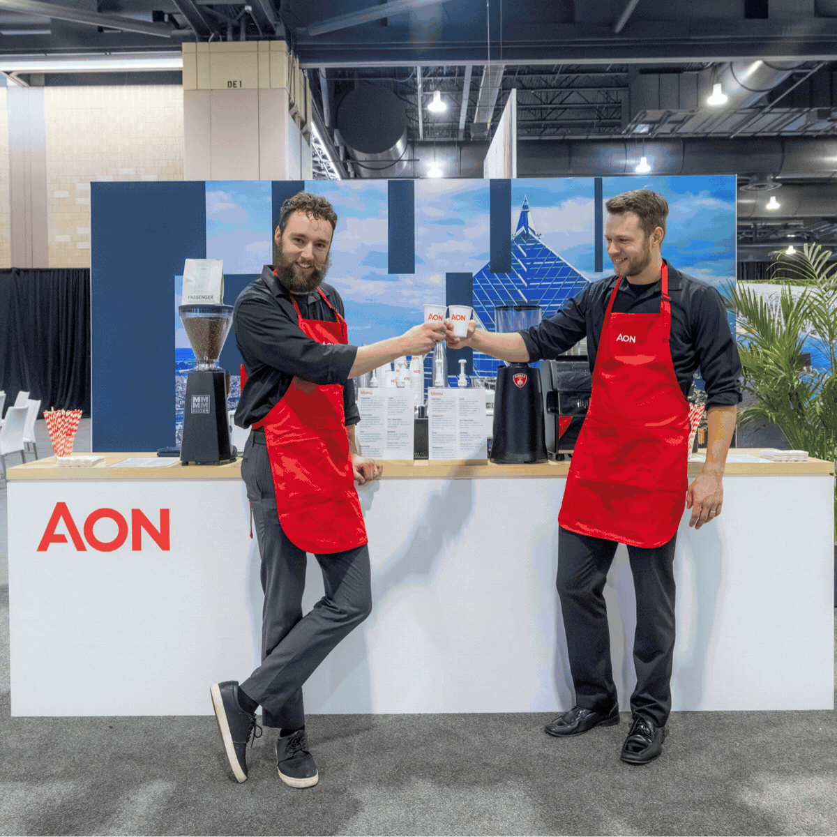 Two men in black shirts and red aprons at a coffee stand exchange coffee cups, with coffee equipment and a blue sky mountain backdrop behind them. The stand features the AON logo.