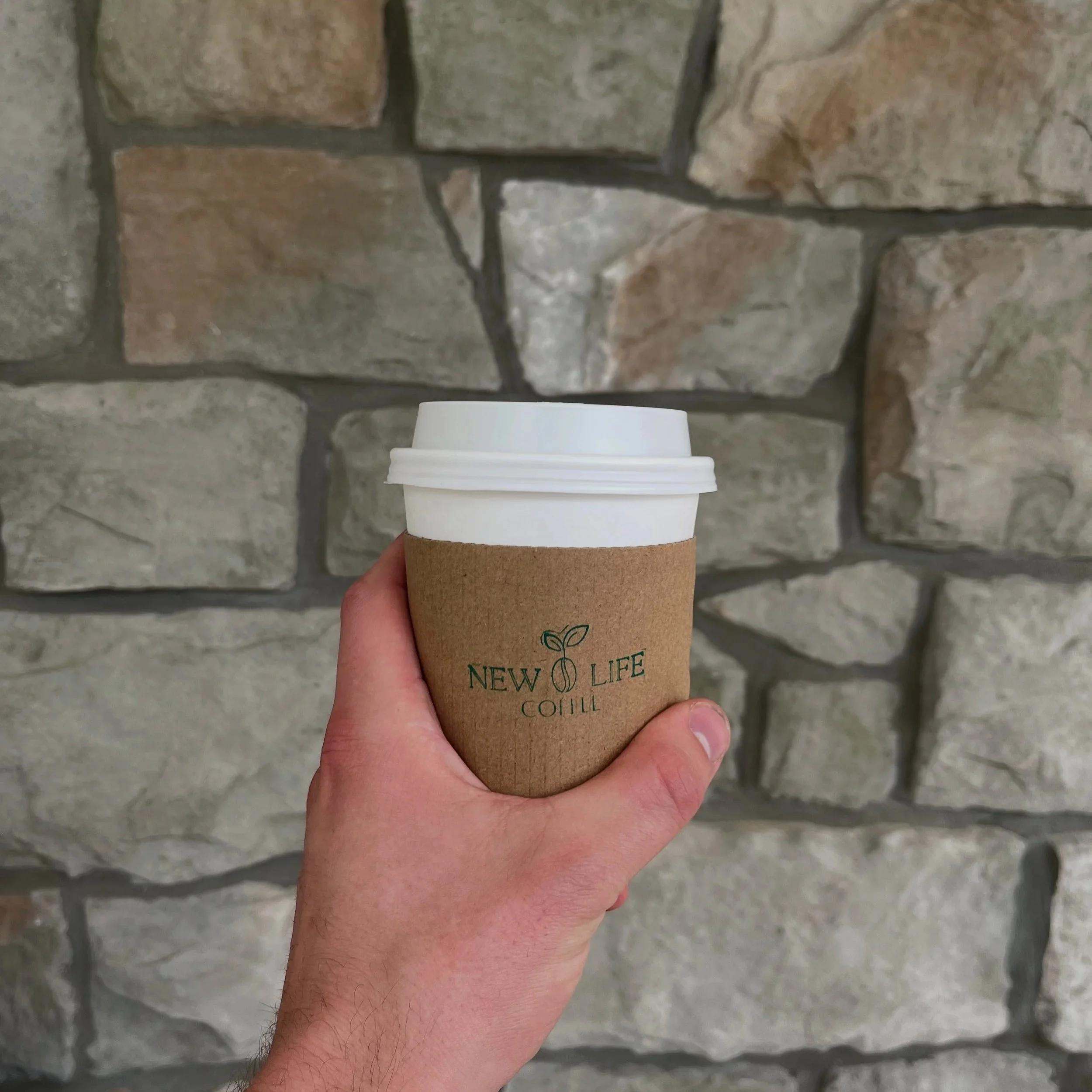 Hand holding a white disposable coffee cup with a brown cardboard sleeve against a stone wall background.