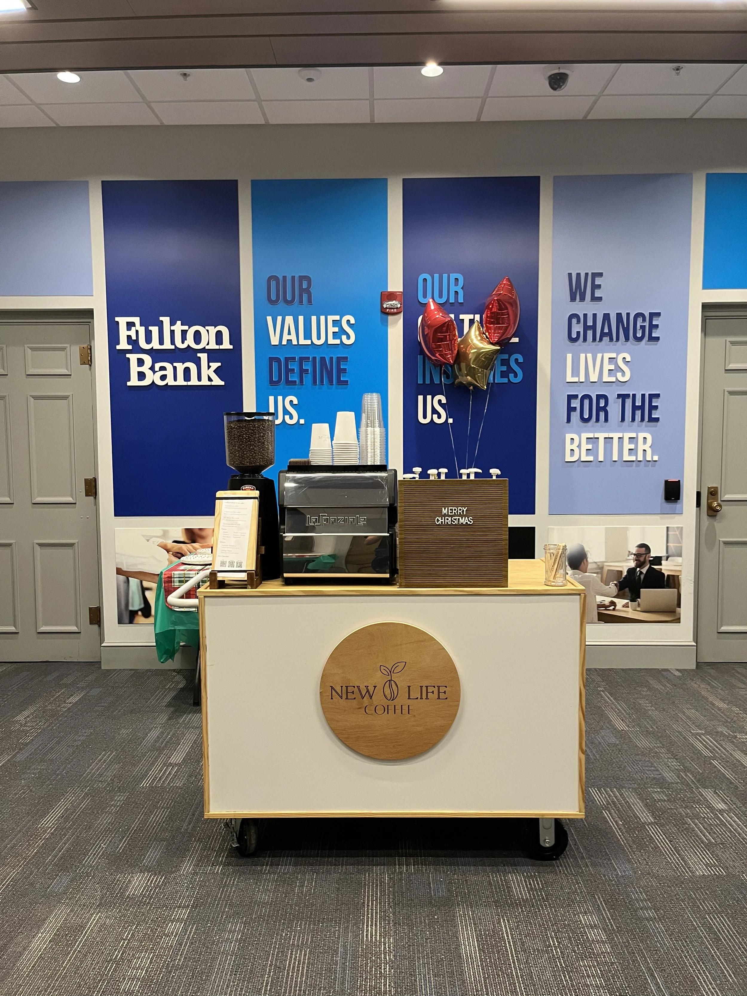 Coffee stand at Fulton Bank with a sign that says 'Our Values Define Us,' and balloons with a Christmas message. The stand has a logo that reads 'New Life Coffee' and a small drink dispenser with coffee beans.