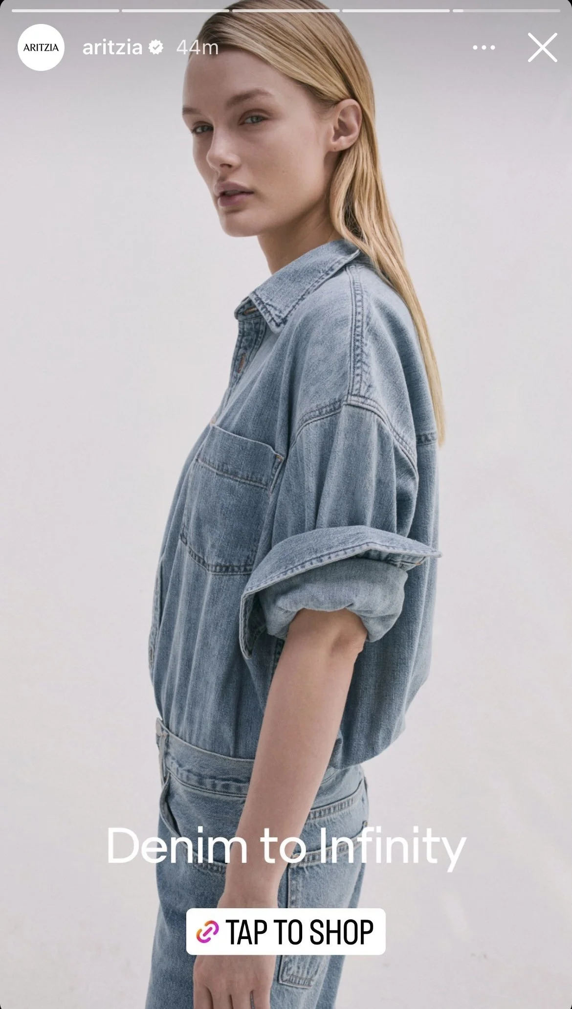 A woman wearing a light blue denim shirt and jeans, standing against a plain white background, looking at the camera with a neutral expression.