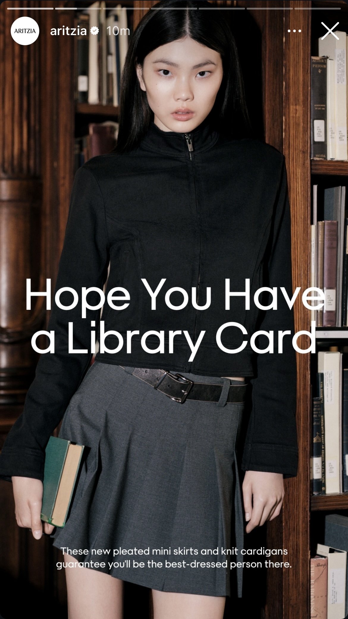 Young woman in a black zip-up jacket and gray pleated mini skirt holding books, standing in front of a bookshelf with a message overlaid that says, 'Hope You Have a Library Card.'