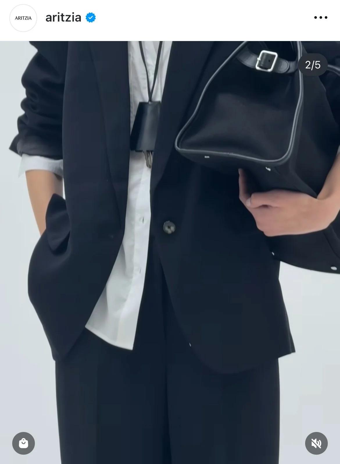 Person wearing a black blazer with rolled-up white shirt sleeves, holding a black bag, and standing against a plain grey background.