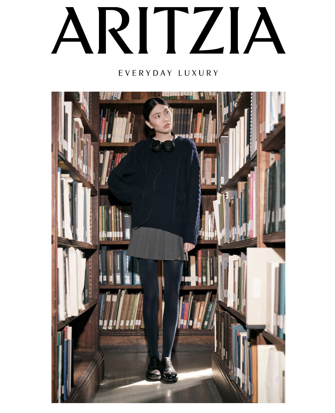 Magazine cover titled 'ARITZIA' with a woman standing between bookshelves in a library, wearing a navy sweater, gray skirt, black tights, black shoes, and headphones around her neck.