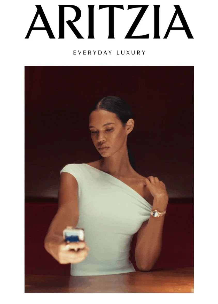 A woman with dark hair pulled back, wearing a white off-the-shoulder top and a watch, looking at her phone. The background is dark red. The image is part of a fashion magazine cover titled 'ARITZIA' with the subtitle 'Everyday Luxury'.